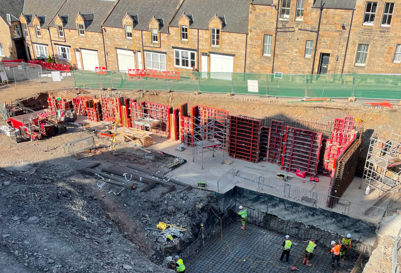 Construction workers are on-site, collaborating on various tasks to build a structure, with safety gear visible.
