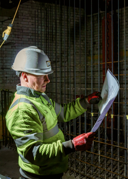 A man in a hard hat and safety vest stands holding a piece of paper, possibly discussing project details or safety protocols.