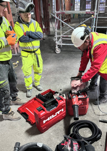 Three men in safety vests and hard hats work together on a red and black HILTI machine at a construction site.